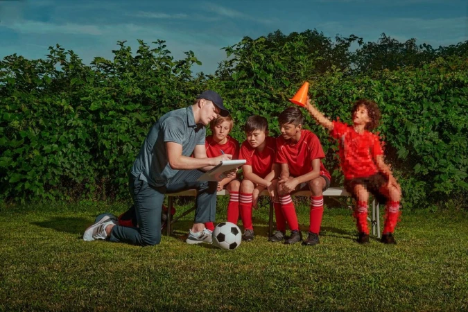 Soccer coach kneels next to his team on a bench, discussing strategy on a clipboard in an outdoor, grassy field.